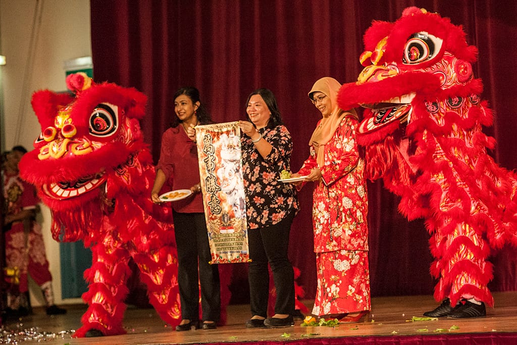 Three women on stage hold a Chinese scroll flanked by two red lion dance costumes during a cultural celebration.